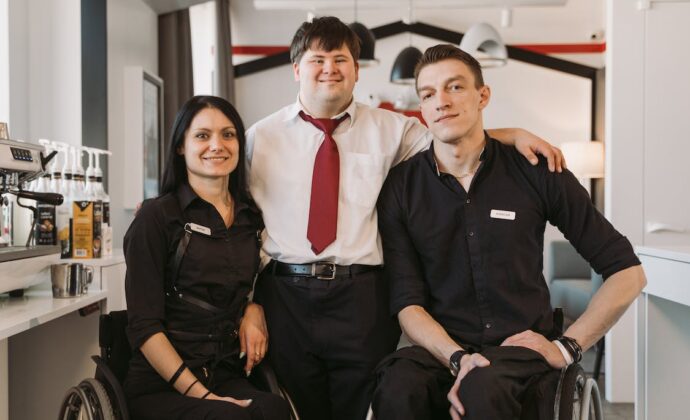 Two people using wheelchairs wearing black work uniforms pose with a man with down syndrome between them who is dressed in work attire. All are smiling for the camera.