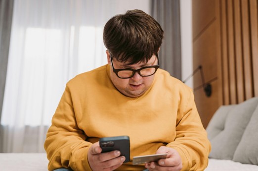 A man with brown hair, black glasses and a yellow shirt sits on his bed looking down at a document and phone he's holding in each hand